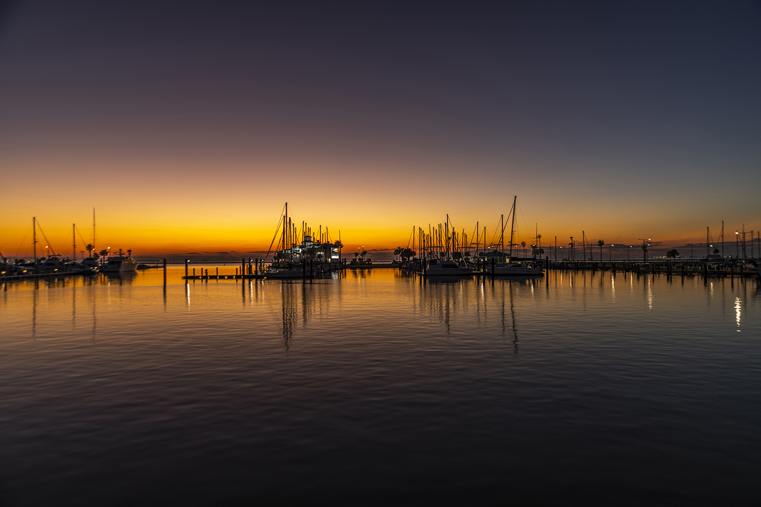 Sailboats docked at a marina in Corpus Christi during sunrise with calm water reflecting the colorful morning sky.