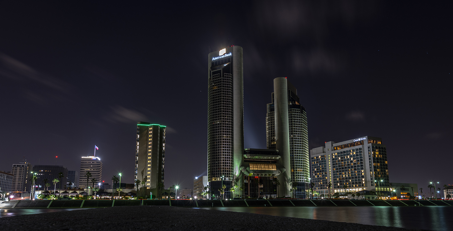 Downtown Corpus Christi skyline at night featuring the American Bank Center tower and illuminated waterfront buildings reflected along Corpus Christi Bay.