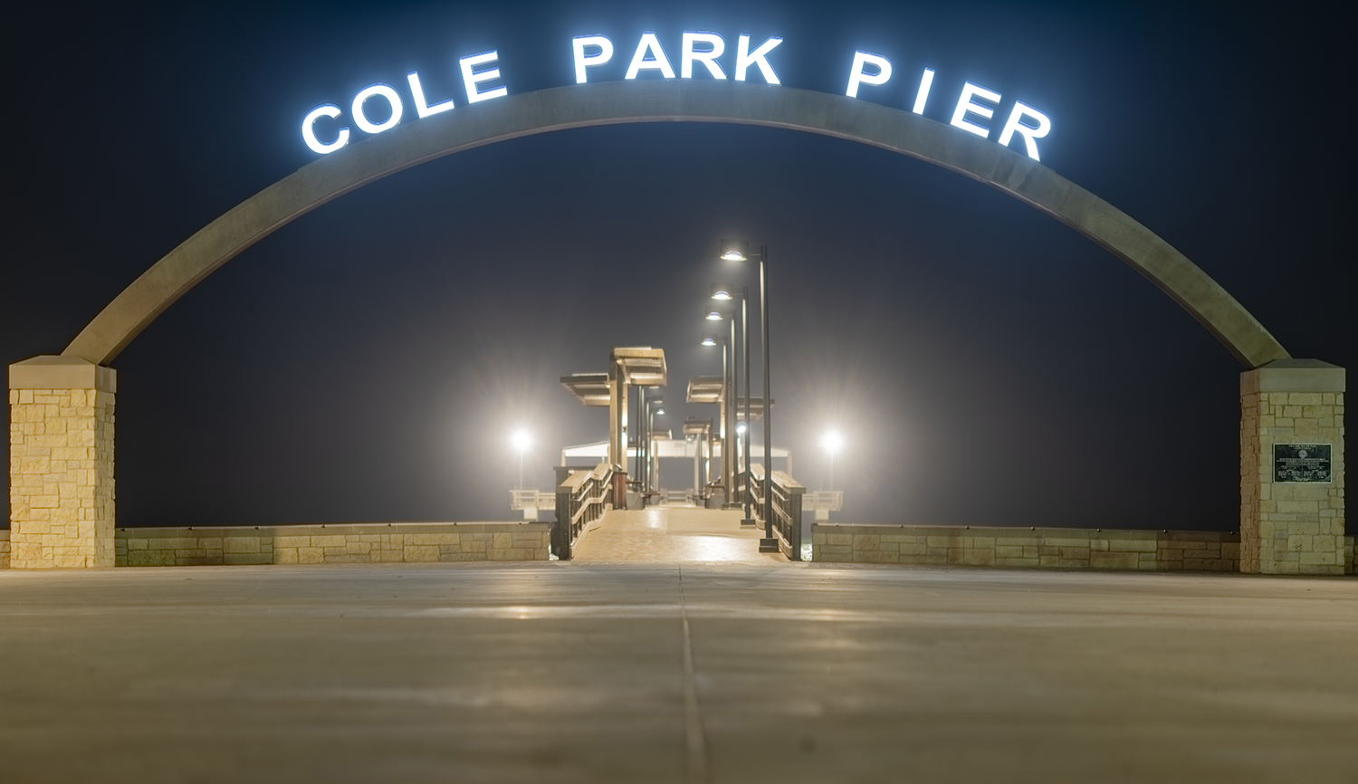 The illuminated entrance arch to Cole Park Pier along the Corpus Christi Bayfront at night.